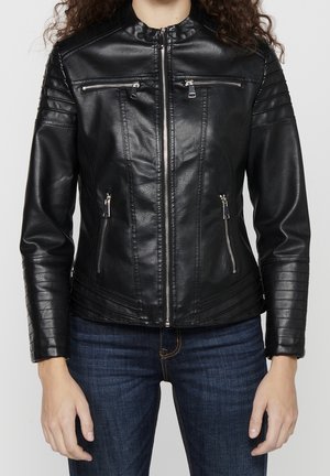 Vrouw draagt een nauwsluitend zwart leren jasje met ritssluitingen en doorgestikte details op mouwen en schouders, gecombineerd met donkerblauwe jeans.