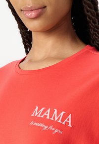 Camiseta de algodón roja con escote en forma de barco y texto bordado en blanco que dice "MAMA te está esperando" en la parte delantera. Textura suave.
