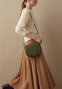Donna con i capelli rossi raccolti, che indossa un maglione beige e una lunga gonna color cammello, mentre porta una borsa a tracolla verde oliva, che cammina davanti a un muro color sabbia.