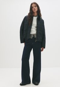 Marinekarierter Blazer mit großen Taschen, kombiniert mit einem grauen Sweatshirt und einem karrierten Hemd, zusammen mit weit geschnittenen dunklen Jeans. Schwarze Schuhe.