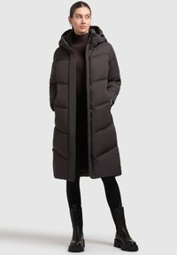 Long black puffer coat with a hood, featuring a quilted design, side pockets, and a straight cut. Worn over a black turtleneck and leggings.