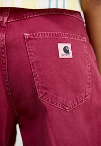 Pantaloni in denim color bordeaux con una tasca posteriore dotata di un'etichetta con il logo. Il tessuto ha una texture liscia ed è cucito con filo bianco a contrasto.