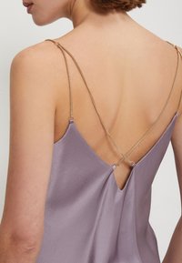 Levandulová saténová košilka s hlubokým výstřihem na zádech, s jemnými zlatými řetízkami, které se kříží ve středu. Hladký povrch.