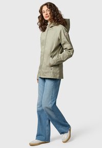 Donna con capelli ricci che indossa una giacca con cappuccio verde chiaro, jeans blu e sneakers bianche, in piedi con una mano in tasca.