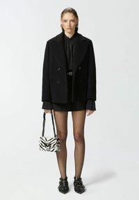 Cappotto doppiopetto nero, blusa nera con maniche a volant, pantaloni corti neri, borsa a righe bianco e nera, tacchi neri con cinturini.