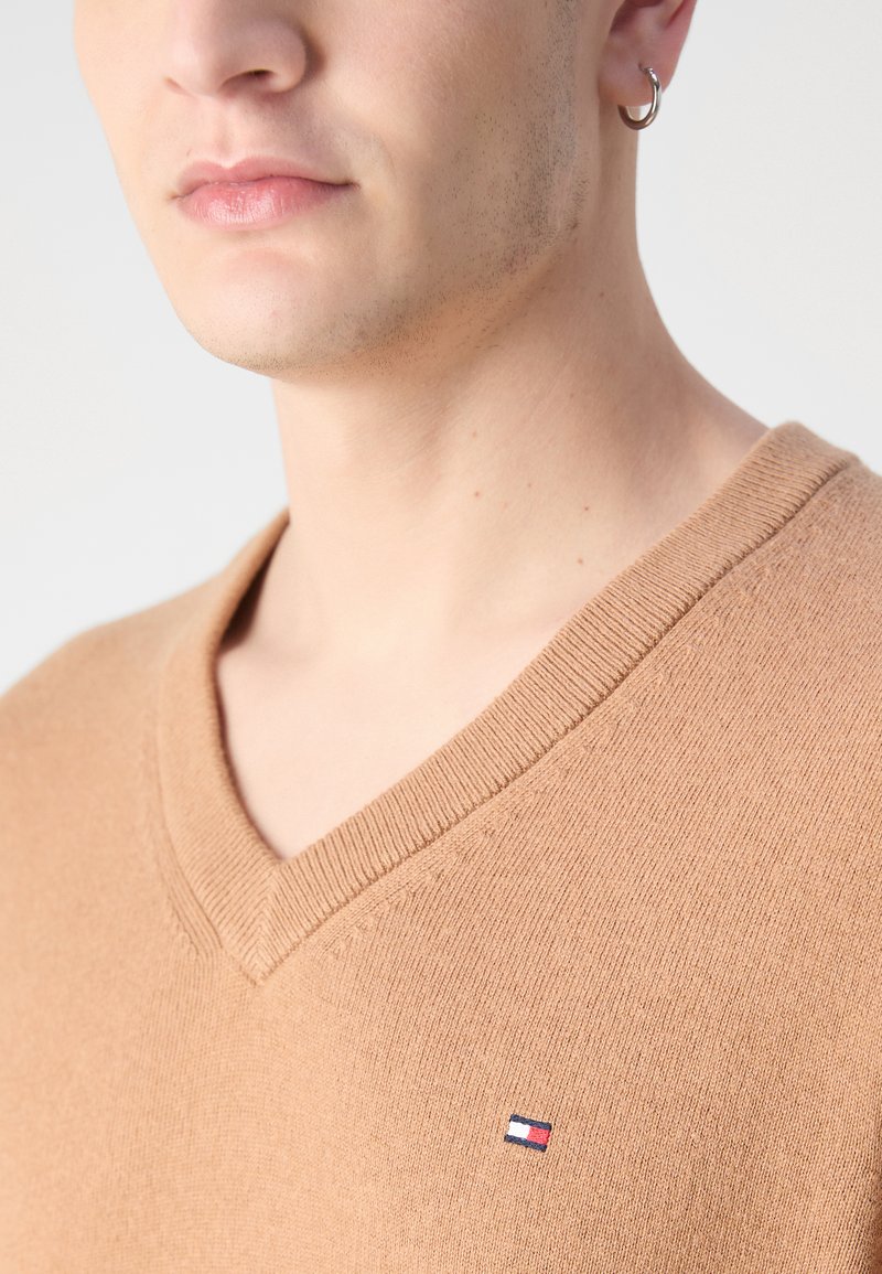 Maglione a v colore marrone chiaro realizzato in morbido tessuto a maglia. Presenta un collo a coste e una piccola etichetta con logo sul petto. Texture liscia.