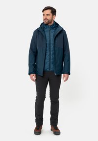 Blauwe geïsoleerde jas met een capuchon, voorzien van een ritssluiting aan de voorkant en zijzakken. Draagt over een lichterblauwe binnenlaag, gecombineerd met zwarte broek.