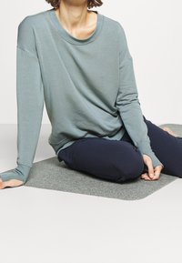 Maglietta a maniche lunghe di un blu chiaro realizzata in un tessuto morbido, abbinata a pantaloni blu scuro. La persona è seduta su un tappetino grigio. Vestibilità rilassata e scollatura tonda.