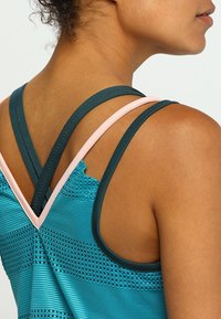 Teal idrottslinne med korsade mörka och ljusa remmar, texturerat tyg med horisontella ränder och perforerade detaljer.