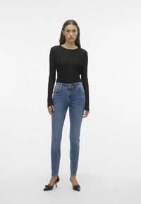 Top negro de manga larga acanalada, ajustado, combinado con jeans ajustados azules con bolsillos frontales, cintura alta y cierre de dos botones. Tacones negros.