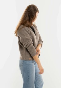 Lichtbruine geribbelde gebreide cardigan met een sjawlcollar en opgestroopte mouwen, gedragen over een zwarte top, gecombineerd met lichtblauwe high-waisted jeans.