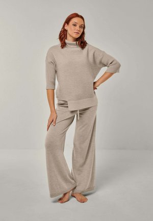 Red-haired woman stands barefoot wearing loose beige ribbed knit sweater with three-quarter sleeves and matching wide-leg pants.
