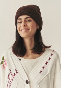 Biały sweter na drutach z haftowanymi kwiatami i różowymi akcentami, noszony na jasnej bluzce, dopełniony brązową dzierganą czapką.