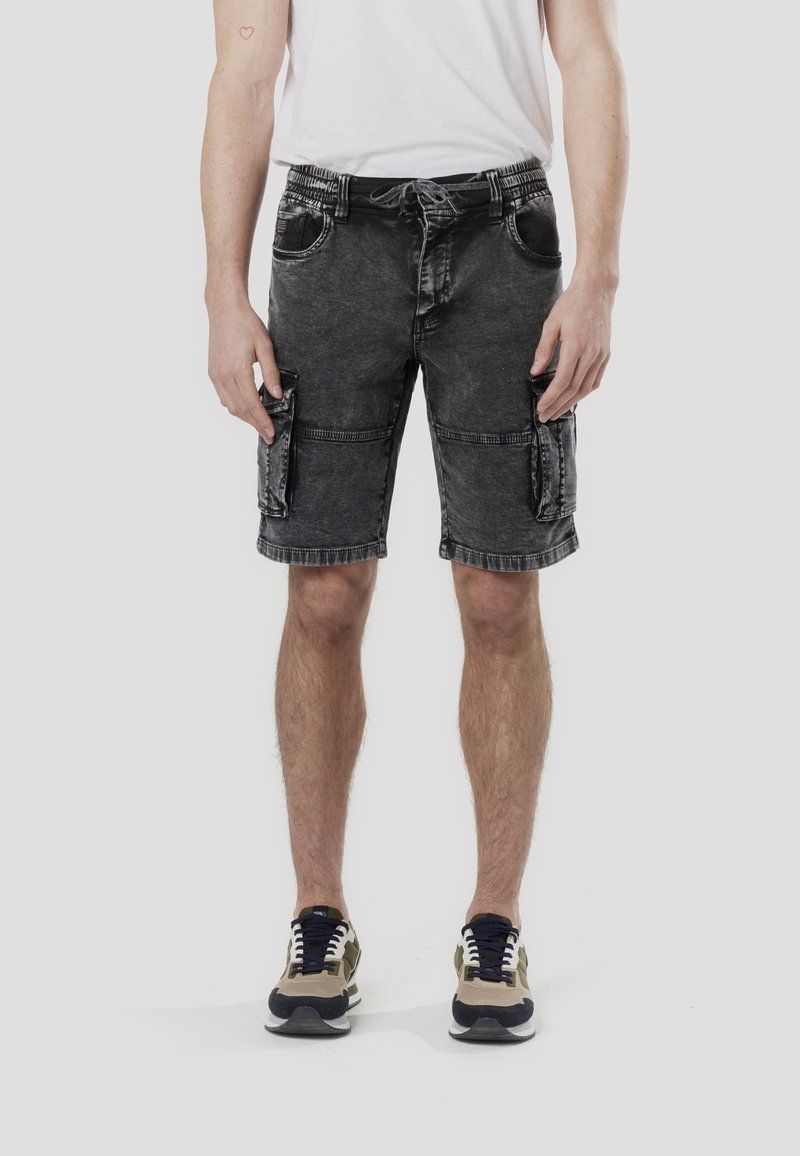 Pantalones cortos cargo de mezclilla negra con cintura elástica, dos bolsillos laterales y dos bolsillos en las piernas. Combinados con una camiseta blanca y zapatillas negras y verdes.