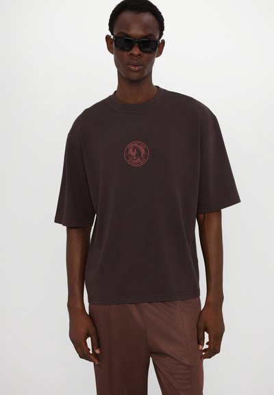 Mørkebrun oversized t-skjorte med korte ermer, med et rødt sirkulært logo trykk på brystet. Glatt bomullstekstur.