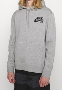 Sudadera gris con capucha que presenta un bolsillo tipo canguro, cordón ajustable y el logo negro de Nike SB en el pecho. Tela suave con puños y dobladillo acanalados.