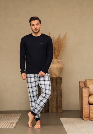Man wearing black long-sleeve shirt and plaid pajama pants stands barefoot in a beige room with rustic decor and leather armchair.