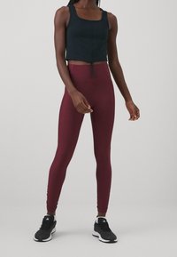 Svart kort topp med rundad halsringning, kombinerad med högmidjade burgundyröda leggings. Modellen bär svarta träningsskor.