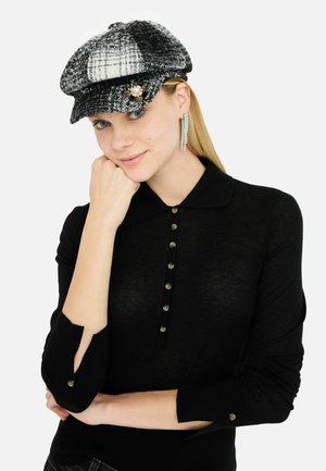 Tweed cap in black and white with a structured shape and decorative brooch. Model wears a fitted black top with a collar and button details.
