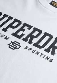 Hvid bomulds T-shirt med fed sorte bogstaver: "SUPERDRY PREMIUM SPORTING." Teksten inkluderer et logodesignelement under ordet "SUPERDRY."