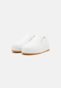 Sapatilhas brancas slip-on feitas de material sintético liso com uma sola de borracha espessa, apresentando um detalhe em castanho. Bico arredondado e design minimalista.