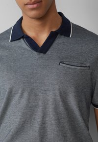 Siva polo majica z mornarsko modrim ovratnikom in belimi pasovi, ki ima majhen dekorativen žep na prsih z vodoravnimi črtami. Mehka tekstura.
