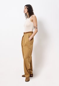 Beige geribbelde tanktop gecombineerd met beige, loszittende, hoge taille broek. Bruin gevlochten riem en sandaaltjes met bandjes. Minimalistische achtergrond.