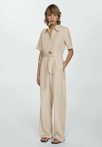 Jumpsuit beige con maniche corte, colletto, cerniera frontale, cintura con fiocco in vita e pantaloni a gamba larga. Presenta tasche laterali. Tessuto liscio.