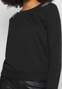 Svart sweatshirt i mjukt tyg, med långa ärmar, rund halsringning och utsmyckningar på axlarna.