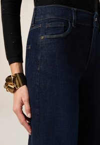 Jeans de mezclilla azul oscuro con una cintura alta, costuras sutiles y bolsillos frontales. Se puede ver una pulsera de oro en la muñeca.