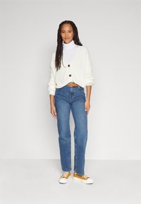 Cream oversized cardigan with large buttons, white turtleneck, blue denim jeans, and yellow sneakers with white soles.