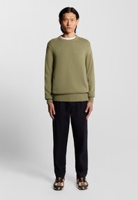 Pull en tricot vert olive avec un col rond, des poignets et un ourlet côtelés. Porté avec un pantalon noir et des chaussures brunes à motifs.