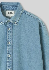 Chemise en denim bleu clair, avec un col boutonné et une patte de boutonnage à l'avant avec des boutons blancs. Texture lisse et design décontracté.