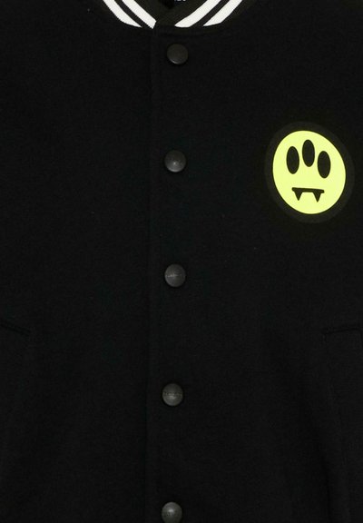 Veste noire boutonnée avec col rayé blanc et patch jaune représentant une patte avec des crocs de vampire sur la poitrine gauche.