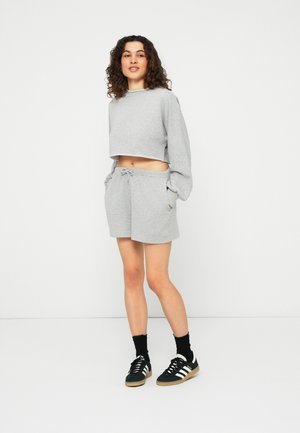 Kvinde står med hænderne i lommerne iført en grå cropped sweatshirt, matchende grå shorts, sorte sokker og sorte Adidas sneakers.