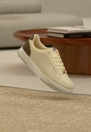 Kremowo-brązowy sneaker z sznurowadłami unoszący się nad teksturowanym beżowym dywanem, w tle drewniany stół i książki.