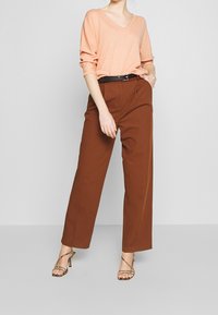 Maglione pesca con scollatura a V e maniche arrotolate, abbinato a pantaloni a gamba larga marroni a vita alta. Cintura nera e tacchi metallici con cinturini.