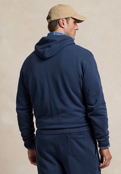 Marineblå hættetrøje lavet af blødt materiale med en kængurulomme, der har en hætte med snoretræk og ribstrikkede manchetter, båret sammen med en beige kasket.
