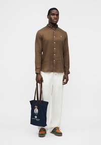 Άνδρας με καφέ πουκάμισο με κουμπιά, λευκό παντελόνι, πολύχρωμα παπούτσια, κρατώντας μπλε τσάντα ώμου με σχέδιο αρκούδας και καφέ δερμάτινες λαβές, στέκεται σε λευκό φόντο.