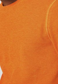 Oranssi poolopaita, valmistettu strukturoitua kangasta, jossa pyöreä kaula-aukko ja kontrastiväriset saumat.