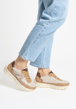 Person trägt hellblaue Jeans und beige Turnschuhe mit weißen Lammfell-Details und dicken weißen Sohlen, steht vor weißem Hintergrund.