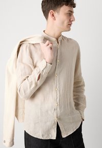 Camicia di lino beige chiaro con colletto alla coreana, chiusura con bottoni e maniche arrotolate. Una giacca di colore chiaro è appoggiata sulla spalla.