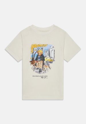 Cremefarbendes Baumwoll-T-Shirt mit einem grafischen Druck eines Bären in einer Jacke, der auf einer Bank sitzt. Bemerkenswerte Elemente sind gelbe Bäume und ein Hund.
