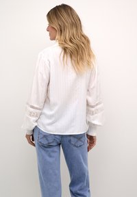 Vit blus med randig textur och spetsdetaljer på ärmarna, kombinerad med ljusblå jeans med unika sömmar på bakfickorna.
