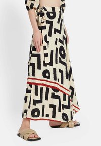 Falda maxi en crema con un patrón abstracto negro y una raya roja. Presenta una cintura atada y un tejido fluido, combinada con sandalias planas en tonos neutros.