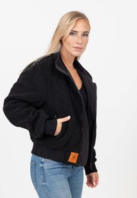 Giacca bomber nera realizzata in pile testurizzato, con zip frontale, polsini a costine, tasche laterali e un'etichetta arancione del marchio sull'orlo.