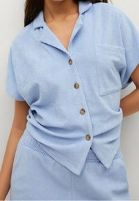 Světle modrá košile s krátkým rukávem vyrobená z měkkého materiálu, se skládacím límcem, zapínáním na knoflíky a jednou náprsní kapsou.