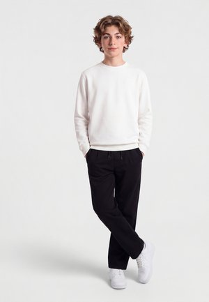 Jeune personne debout, les mains dans les poches, portant une chemise blanche à manches longues, un pantalon noir et des baskets blanches sur un fond uni.