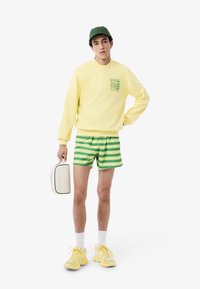 Geel sweatshirt met groen logo, groen en witte gestreepte shorts, witte tas en gele sneakers; korte mouwen en een comfortabele pasvorm.