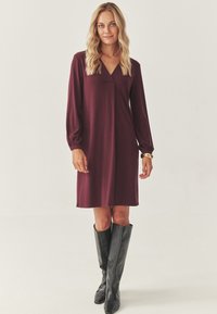 Femeie blondă îmbrăcată într-o rochie burgundă cu mâneci lungi, până la genunchi și cizme negre până la genunchi, stând pe un fundal alb simplu.